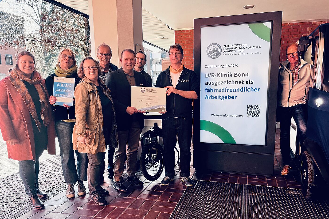 Zertifikatsübergabe LVR-Klinik Bonn Fahrradfeundlicher Arbeitgeber in Silber Mehrere Menschen stehen links und rechts verteilt um einen vertikalen Screen, auf dem steht "LVR-Klinik Bonn ausgezeichnet als fahrradfreundlicher Arbeitgeber".