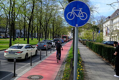 Ein geschützter Radfahrstreifen mit Roteinfärbung. Radwegschild und einer Radfahrerin. 