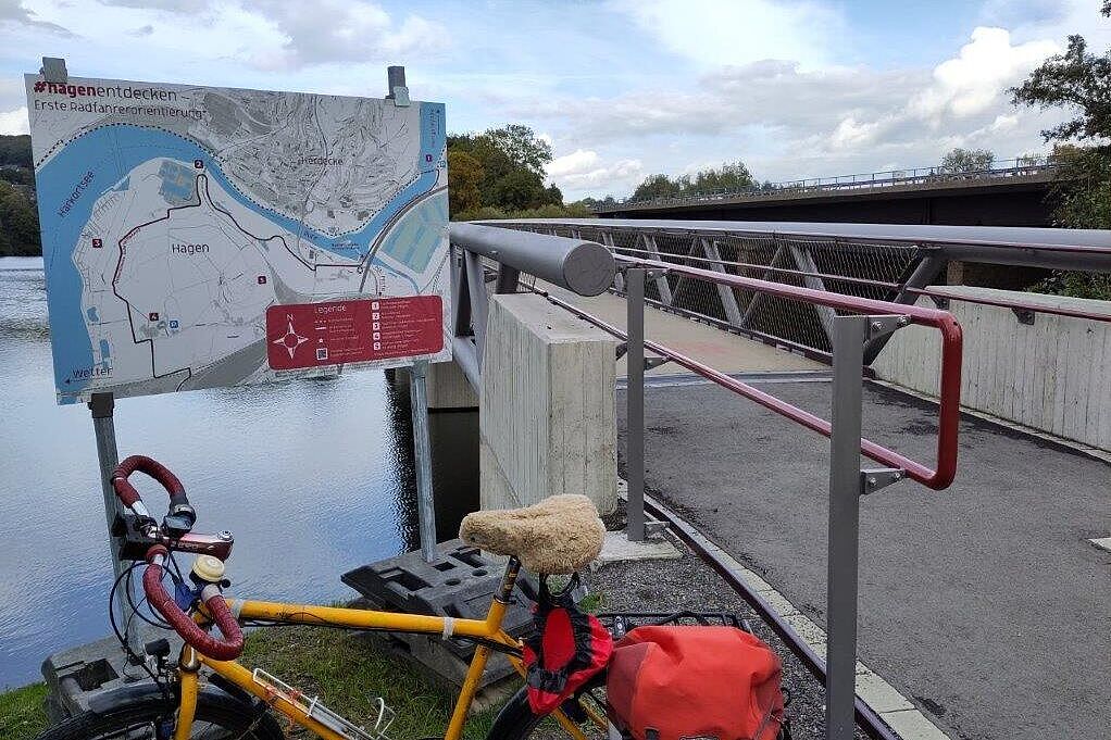 Orangenes Fahrrad mit roter Gepäcktasche steht vor Karte. Im Hintergrund Fluss und Brücke.