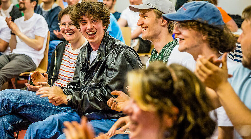 Viele Menschen sitzen in einem Raum, klatschen und lachen. Die Menschen sehen jung aus. In der Bildmitte sitzt ein junger Mann in Lederjacke und mit lockigen Haaren. 