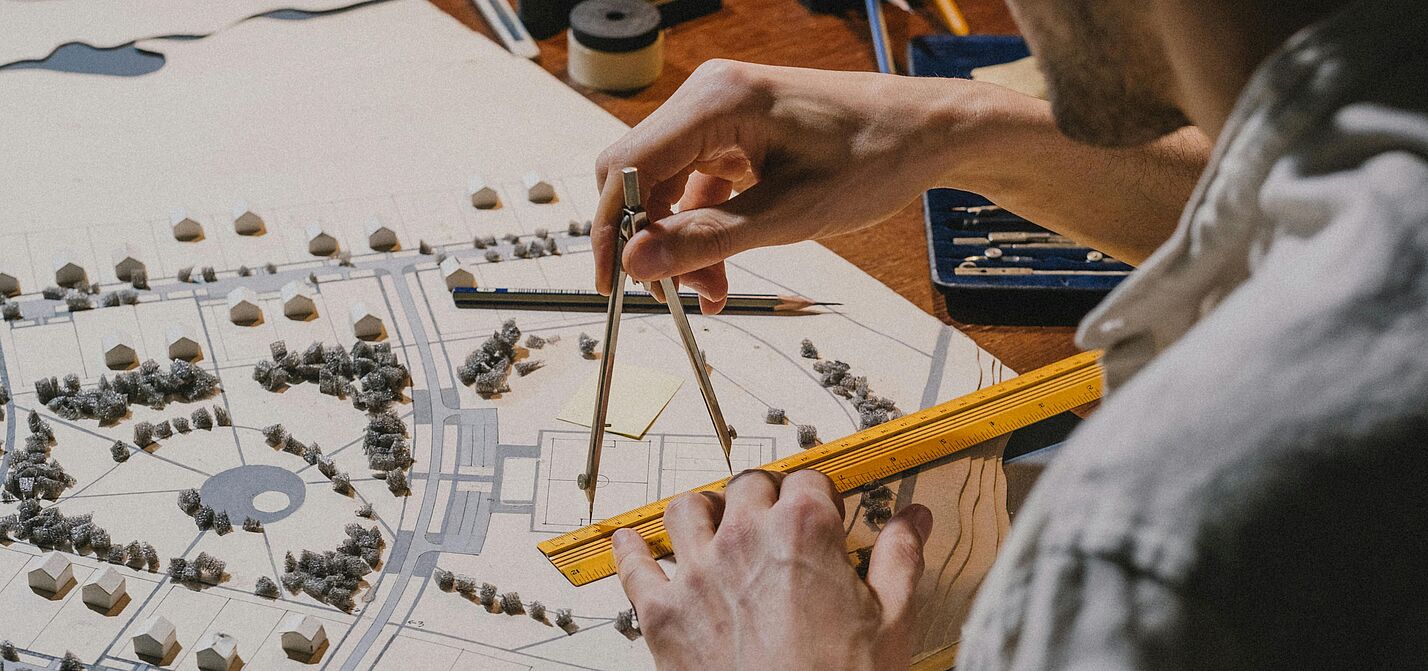 Ein Mann im Hemd ist von hinten zu sehen und zeichnet mit Lineal und Zirkel etwas auf einen Stadtplan.
