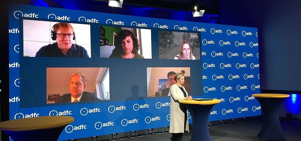 InnoRAD-Symposium Ein Studio mit der Moderatorin am Pult, dahinter eine Videowand mit den Köpfen von per Video zugeschalteten Personen.
