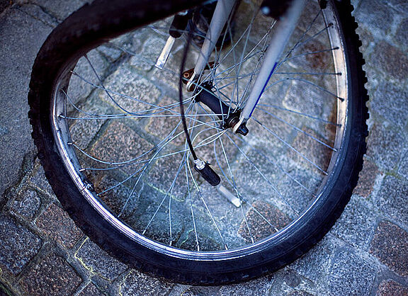 Verbogene Fahrradfelge Nahaufnahme eines Fahrradvorderrads auf Kopfsteinpflaster. Die Felge ist verbogen und mehrere Speichen sind gebrochen oder hängen lose herab. Das Rad steht schief auf dem grauen Pflaster und ist offensichtlich nach einem Unfall nicht mehr fahrbereit.