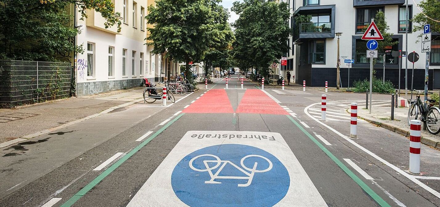 Fahrradstraße Berlin Eine Straße in Berlin: Rechts und links sind Häuserzeilen zu sehen, die Fahrbahn ist mit einem großen Fahrradsymbol versehen. Weiter hinten ist die Fahrbahn rot eingefärbt, Poller dienen als modaler Filter.
