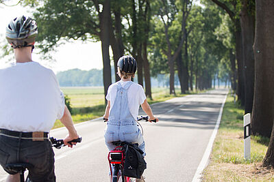 Zwei Personen mit Fahrradhelmen fahren auf einer schmalen Landstraße ohne separaten Radweg.